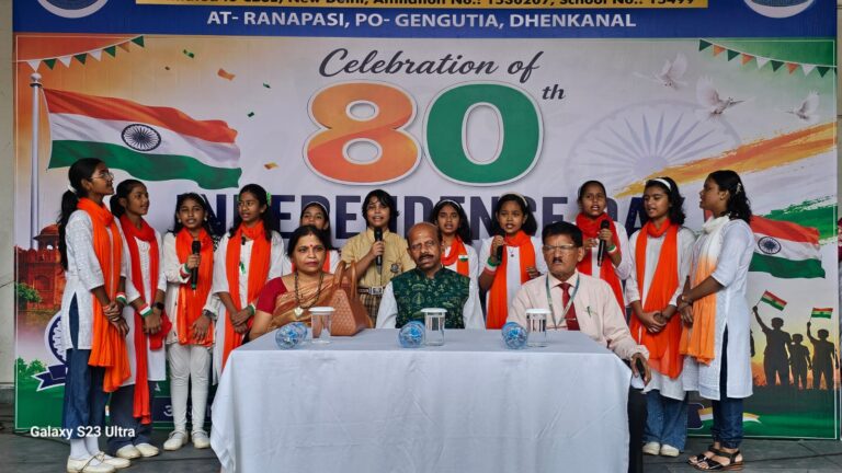 Schoolgirls in white outfits with orange stoles sing on stage beside a table of officials during an 80th Independence Day celebration banner in the background with the Indian flag.
